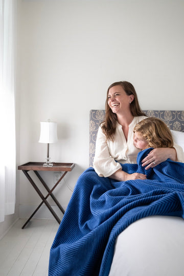 Blue herringbone-patterned blanket draped over a bed with an adult and child sitting on it.2