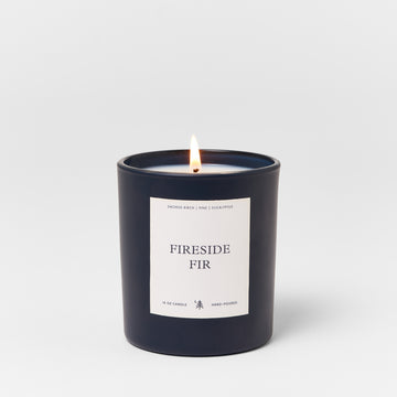 Black cylindrical candle with white label reading "Fireside Fir" and lit flame.4