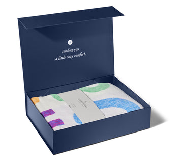 Dark blue gift box with lid open, containing folded white textiles with colorful accents in green, blue, purple, and orange.