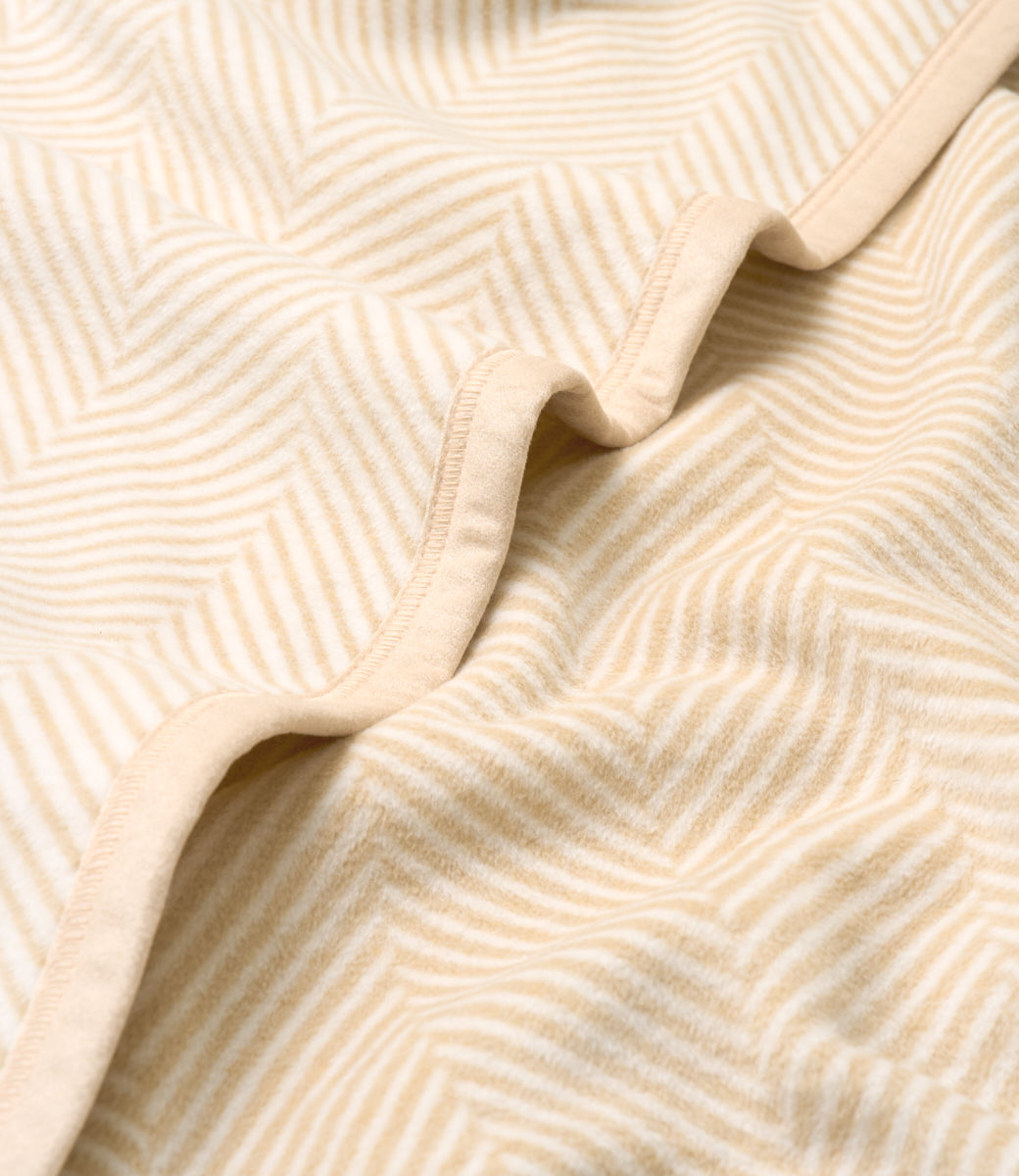 Cream blanket with herringbone pattern and bound edges, shown folded to display texture and weave detail.