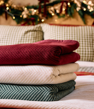 Folded cranberry herringbone midi blanket stacked with cream and green striped blankets on a bed.3