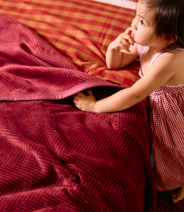 Cranberry and gold herringbone patterned midi blanket draped over a bed with a toddler beside it.5