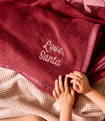 Cranberry red blanket with herringbone texture and white "Love Santa" embroidery, with child's hands resting on it.6