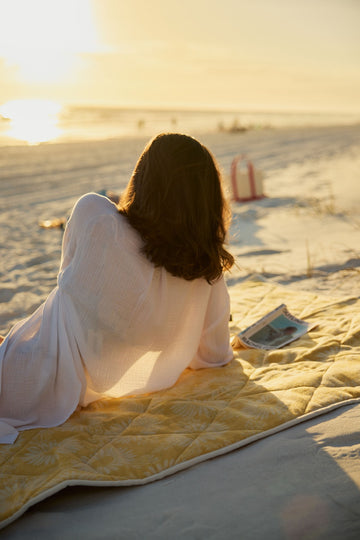 Yellow patterned outdoor blanket spread on beach sand with person sitting on it at sunset.3