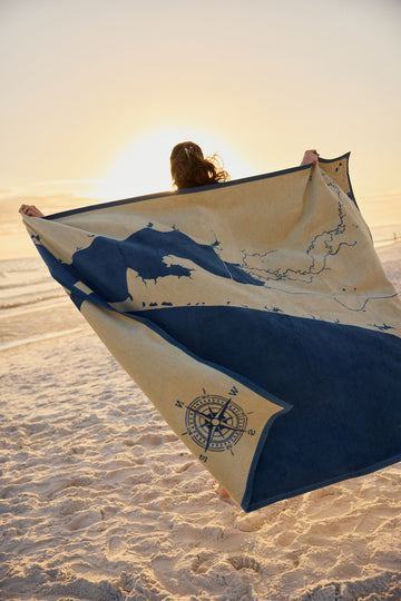 Cream and navy blanket with map design and compass rose held open on beach at sunset.3