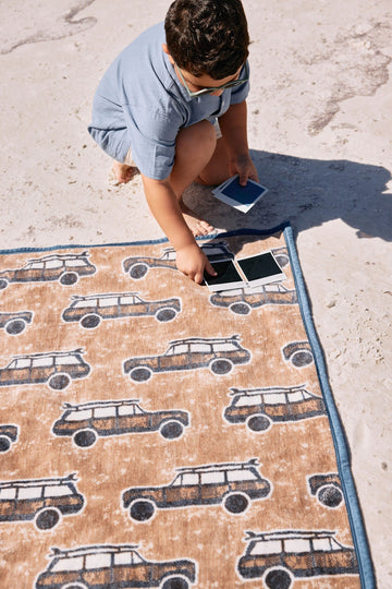 Tan and navy blanket with repeating surf wagon pattern and blue binding, displayed on sand.4