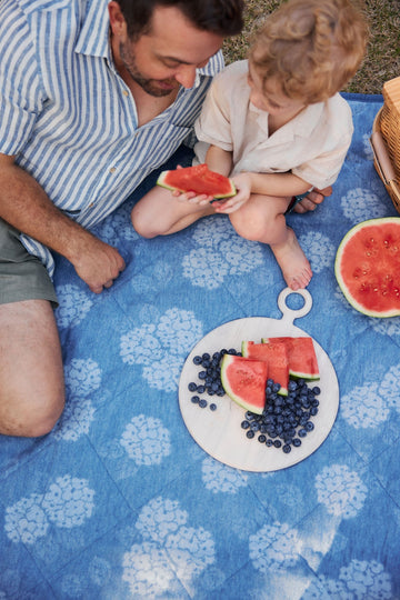 Blue outdoor blanket with white floral pattern spread on grass, adult and child sitting on it with watermelon and berries.5