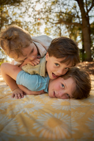 Three children playing on a tan and white patterned outdoor blanket beneath trees.6