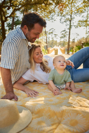 Yellow and cream patterned outdoor blanket spread on ground with family of three sitting on it in shaded park setting.7