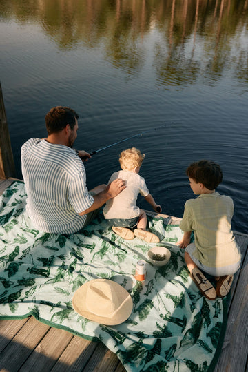 Cream blanket with green tree pattern spread on dock with adult and two children fishing by water.6