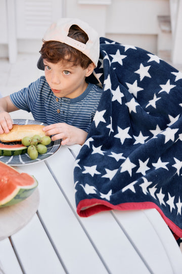 Navy blanket with white stars and red stripe draped over child eating at outdoor table.9