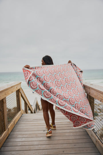 Blanket with red and teal painted shell pattern held by person on beach boardwalk.3