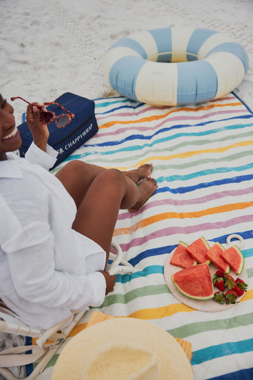 Striped beach blanket in multicolor wavy lines with person sitting on sand holding sunglasses and watermelon slices nearby.3