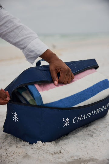 Rolled blanket with colorful stripes in navy, pink, and blue being held open at the beach, with dark blue carry case labeled 7