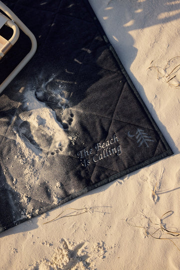Black outdoor blanket with "The Beach is Calling" text, partially covered in sand on a beach next to a white cooler.3