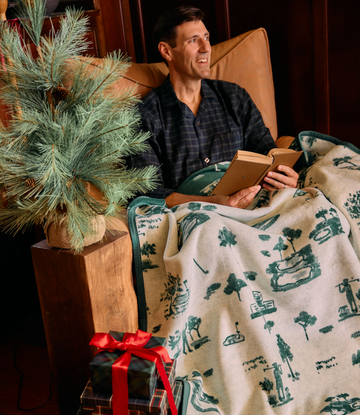 Cream blanket with green tree and nature-themed pattern draped over person sitting in tan chair.2