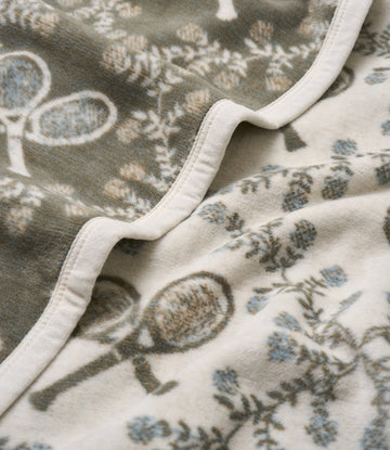 Folded blanket with cream binding showing tennis racket and floral pattern in gray, tan, and blue tones.5