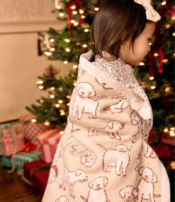 Cream-colored mini blanket with red Santa hat puppies pattern, worn by child in front of lit Christmas tree.2