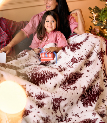 Cream blanket with burgundy toile pattern featuring Santa, reindeer, and snowflakes, displayed on a bed with two people.5