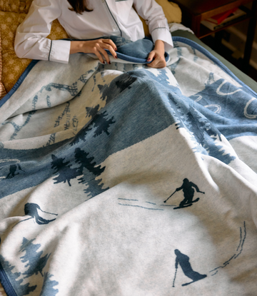 Cream and blue blanket with skiers, snow-covered trees, and mountains printed throughout, displayed on a person's lap.2