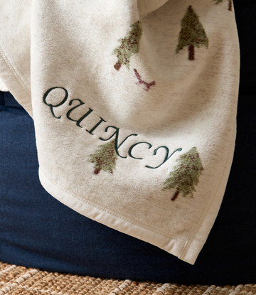 Cream blanket with evergreen tree pattern and text embroidery displayed over dark blue fabric.6