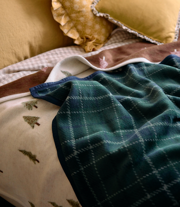 Folded plaid blanket in green and navy with cream lining, displayed on cushions with small tree prints visible.3