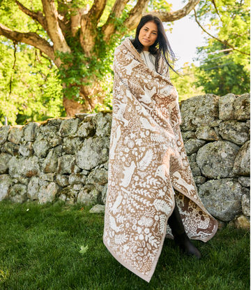 Woman wrapped in tan and cream blanket with woodland animal and botanical print standing against stone wall under tree.2