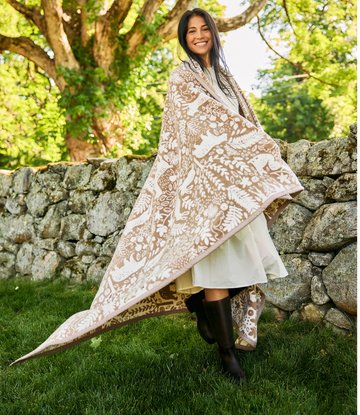 Woman wearing cream and tan blanket with forest-inspired pattern draped over shoulders outdoors against stone wall.3