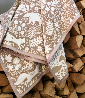 Tan and cream blanket with woodland animal and botanical print, pink trim, draped over stacked logs.4