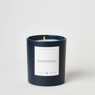 Dark blue glass candle with white label reading "Nightfall," lit flame visible, labeled 10 oz hand-poured candle.3