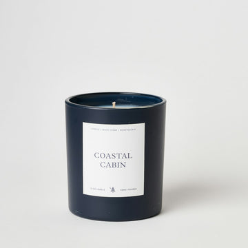 Dark blue cylindrical candle with white label reading "Coastal Cabin" and single wick visible at top.3