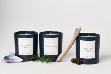 Dark navy cylindrical candle labeled "Nightfall" with white label, displayed among two other scented candles, driftwood, shel4