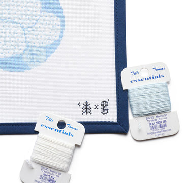 Needlepoint canvas with blue hydrangea design and two spools of Tille Tomas Essentials thread in white and light blue.4