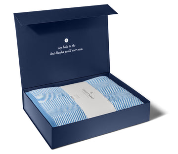 Navy blue gift box with open lid containing blue and white striped blanket and white band label.